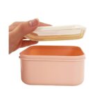 Pink rectangular lunch box being opened by a hand, showing an orange interior and clear lid peel.
